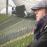 Man staring into an empty football stadium holding a mobile phone with a cap and coat on -Munich, Olympic Stadium, 2020