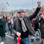 Brendan Ogle holding an umbrella with left fist raised and a crowd behind him at a dispersing protest